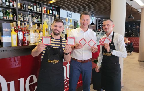 Bürgermeister Jörg Zietemann (Mitte) und die Gebrüder Mustafa, Geschäftsführer des Restaurants „Due Fratelli“ in Rathenow, präsentieren die neuen Veranstaltungsbierdeckel der Stadt Rathenow 